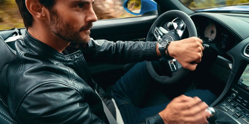 A man in a leather jacket is driving a 2024 Porsche 718 Cayman, holding the steering wheel with both hands