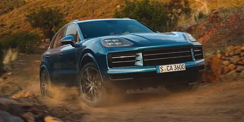 A blue 2024 Porsche Cayenne is driving on a dirt road, creating a cloud of dust behind it. The car appears to be in motion, and the image captures the excitement and thrill of driving a high-performance vehicle on an off-road terrain. The dusty road and the car's speed evoke a sense of adventure and freedom, making the scene visually appealing and engaging.